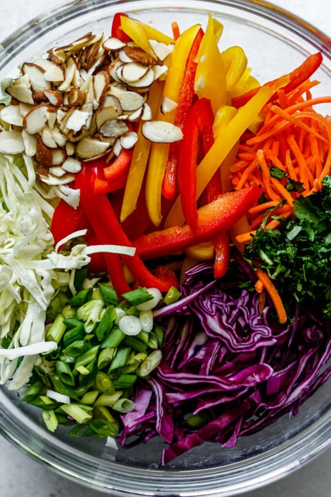 sliced almonds, bell peppers, cilantro, green cabbage, red cabbage, shredded carrots, cilantro, and green onion in a clear bowl