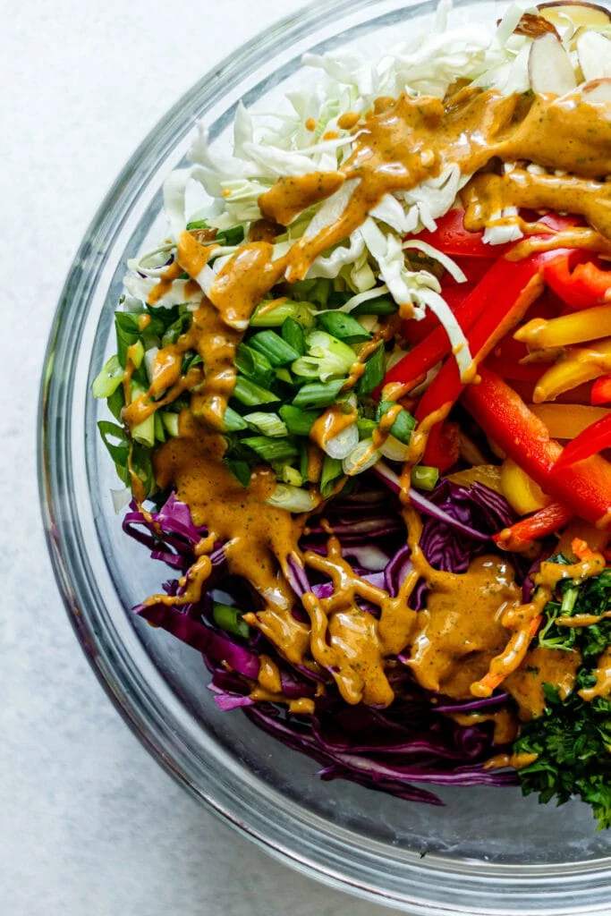 cabbage, bell peppers, cilantro, almonds, and green onions in a clear bowl with peanut dressing drizzled on top