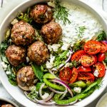 greek meatballs in a white bowl with tzatziki sauce, tomatoes, fresh herbs, green peppers, and onions