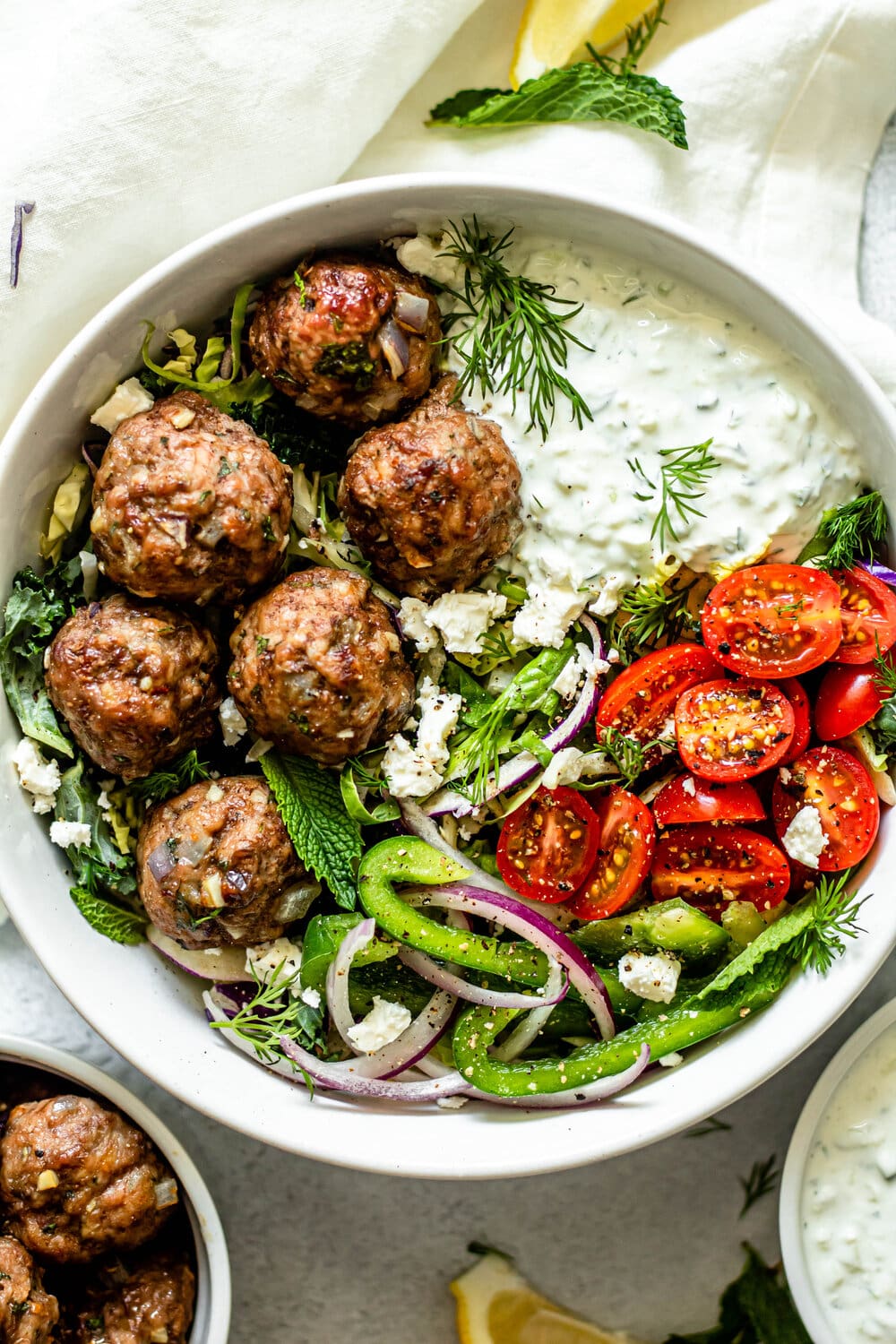 greek meatballs in a white bowl with tzatziki sauce, tomatoes, fresh herbs, green peppers, and onions