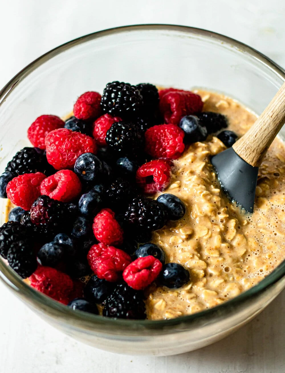 Triple Berry Baked Oatmeal cup batter with fresh berries and wooden spatula