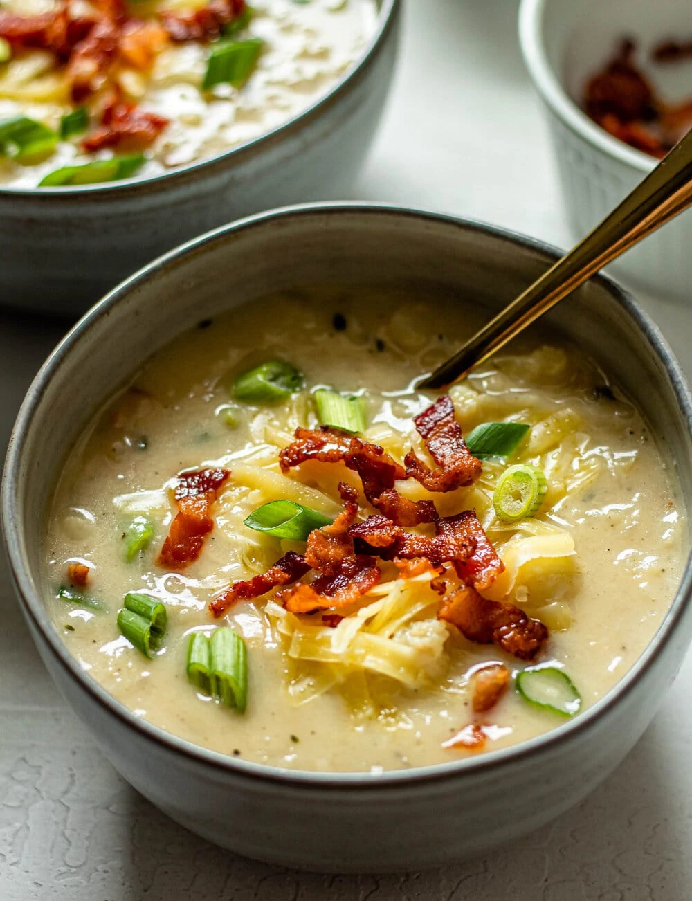 Loaded Cauliflower Soup topped with bacon, green onion, and shredded cheese with a gold spoon on the side