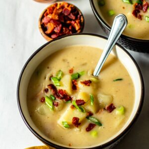 loaded baked potato soup in a brown bowl with a silver spoon and bacon and green onions sprinkled on top