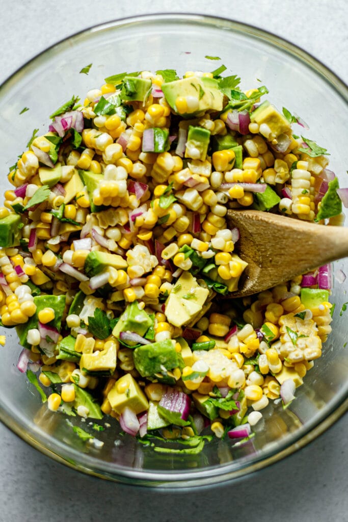 avocado corn salsa in a clear mixing bowl with wooden spoon