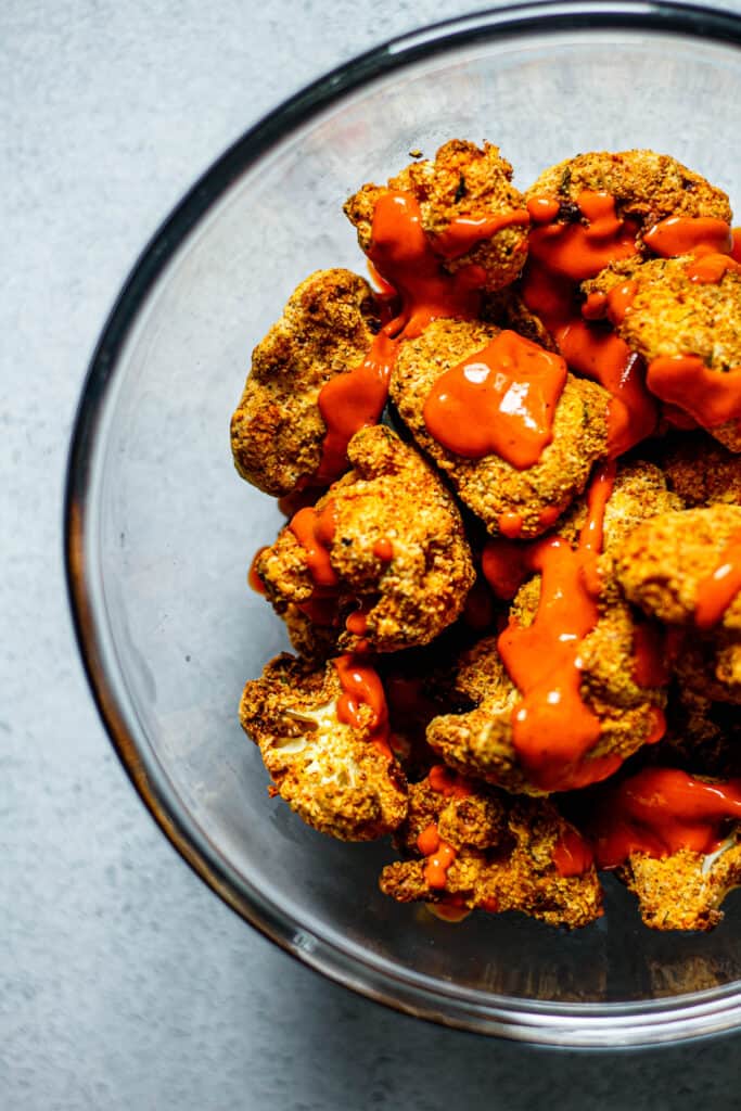 crispy buffalo cauliflower in a clear mixing bowl coated drizzled with buffalo sauce