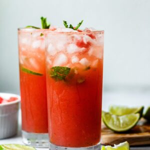 Watermelon Mojito Mocktail in two tall glasses with watermelon and limes in the background