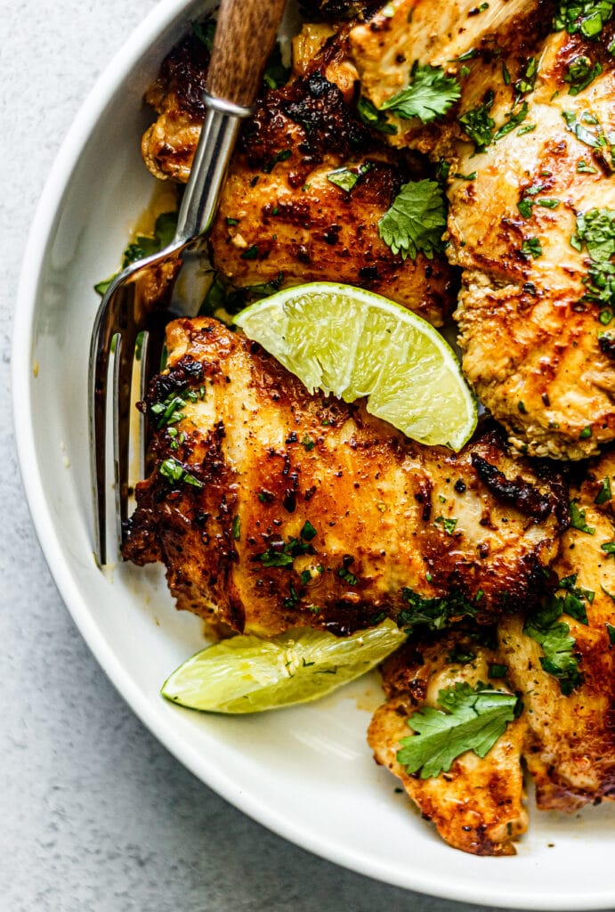 Cilantro lime chicken thighs on a white plate with sliced limes, topped with cilantro, and a fork on the side of the plate