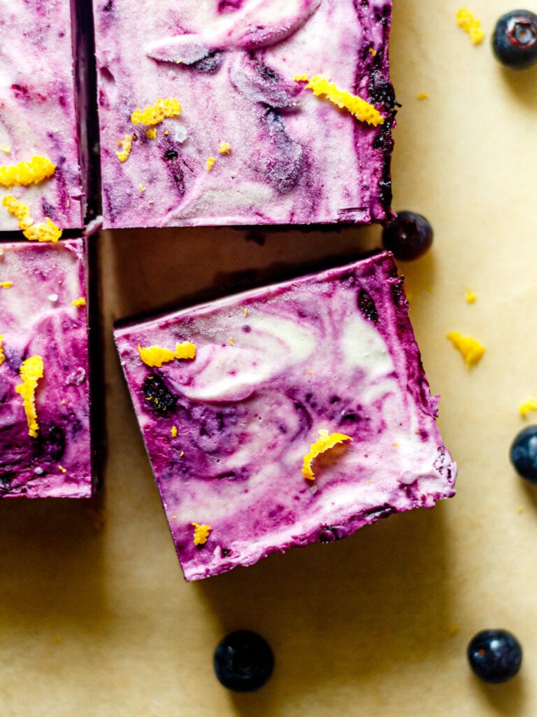 no bake lemon blueberry cheesecake bar on a brown background with lemon zest and blueberries