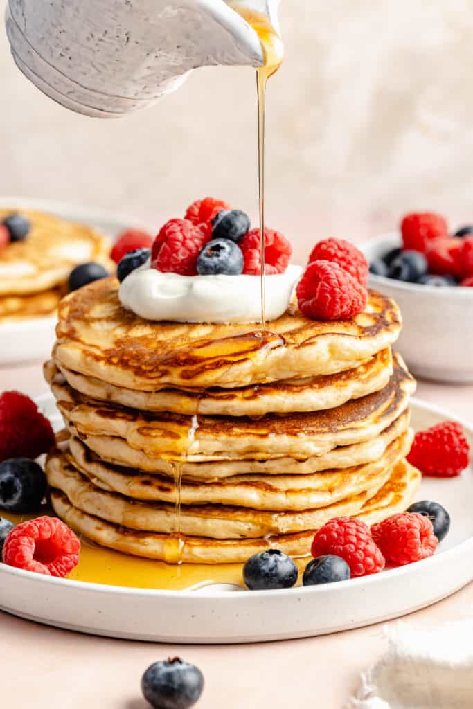 stack of pancakes with cream and berries and maple syrup being drizzled on top