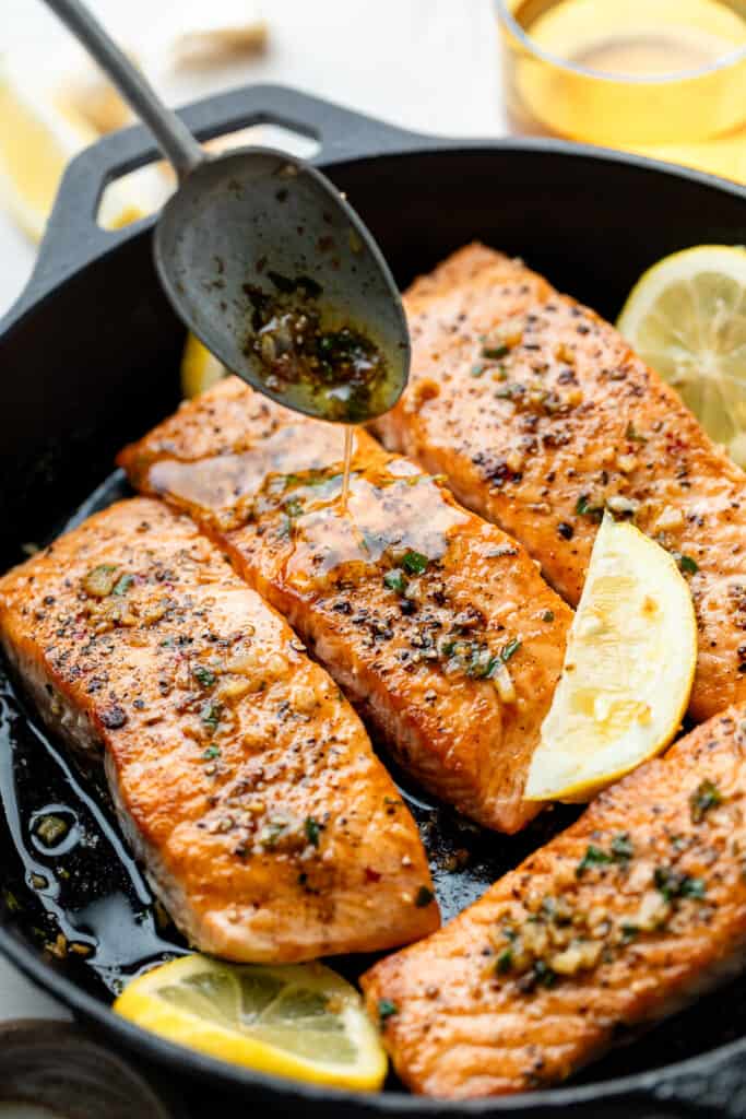 butter sauce being spooned over salmon in cast iron skillet with lemon