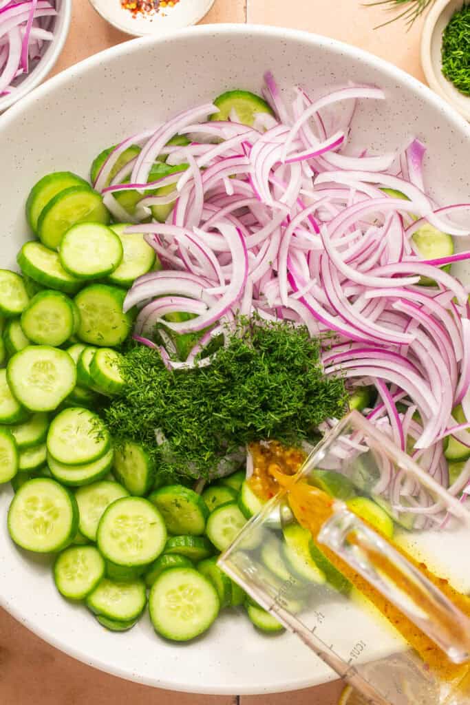 dressing being poured over cucumber salad ingredients in large mixing bowl