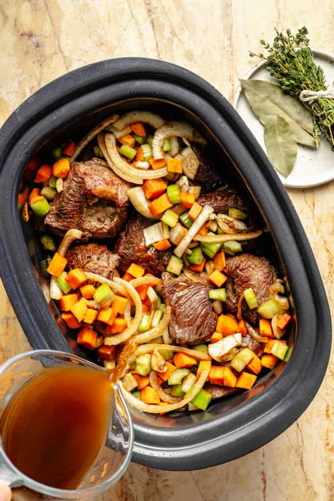short ribs and veggies in slow cooker and broth being poured into slow cooker